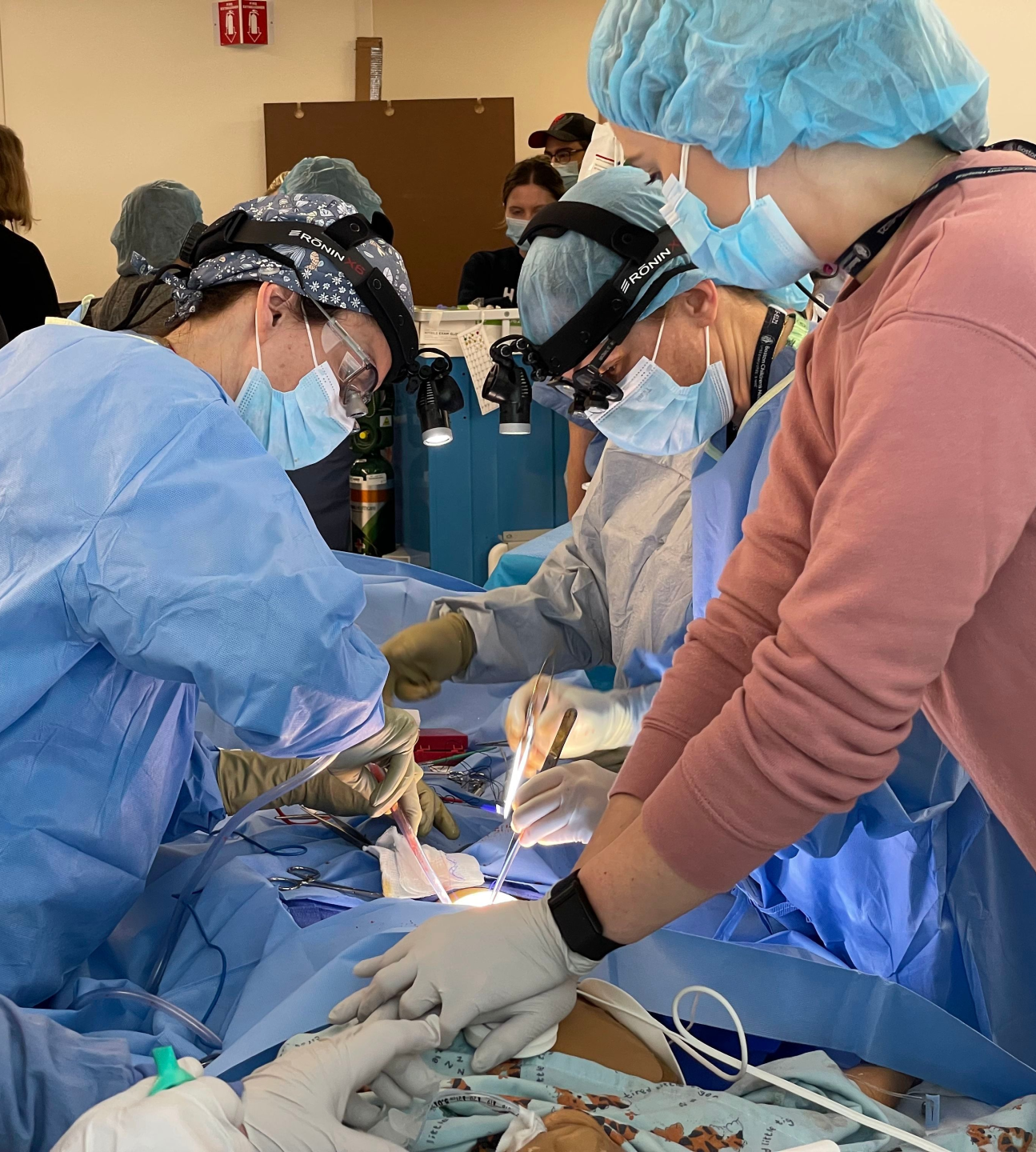 Two doctor in blue scrubs, surgical face masks, and a trainee in a pink sweatshirt practice an operation on a simulation.