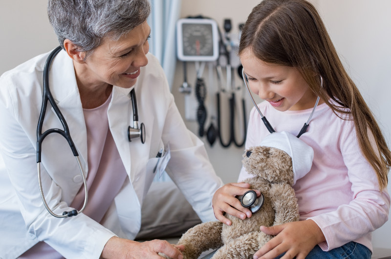 a girl listening to a teddy bear's heart with a stethoscope while a pediatrician looks on