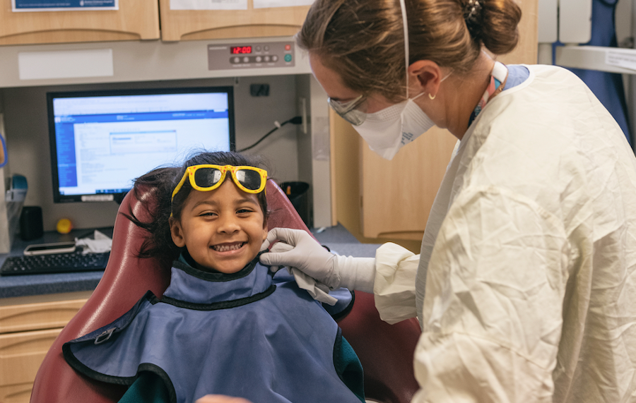 Girl wears protective glasses and smiles as clinician works on her teeth
