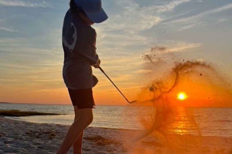 Child swings at golf ball while on beach during sunrise