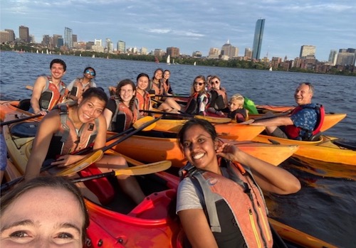 Group of over ten people in kayaks on the Charles River with Boston skyline behind.