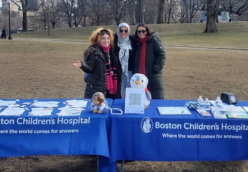Three women in the park standing behind two tables with blue Boston Children's Hospital table cloths