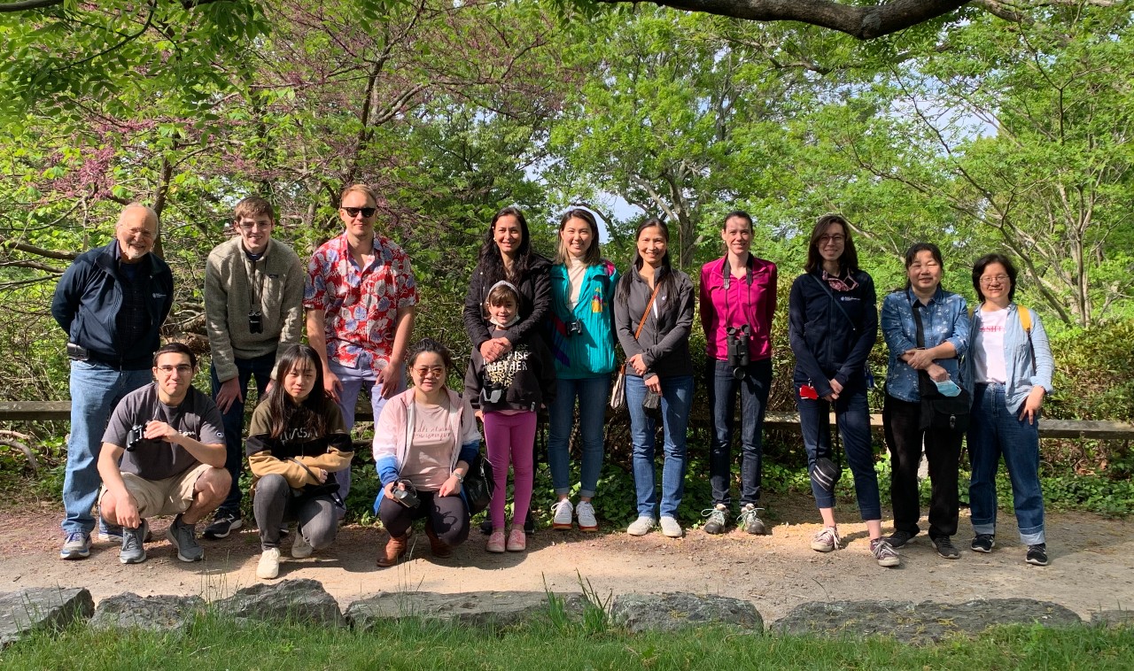 Members of the Benowitz lab all stand together outside under green trees for a team photo.