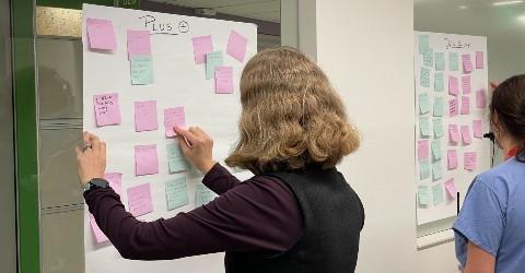 A woman in a long sleeve purple shirt has her back to the camera as she writes on a pink post-it note that's on a white board.