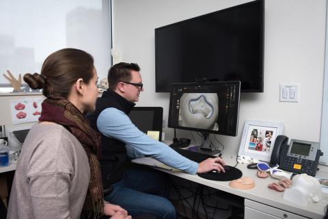 A man and a woman sit at a computer monitor designing a piece of skull to 3D print.
